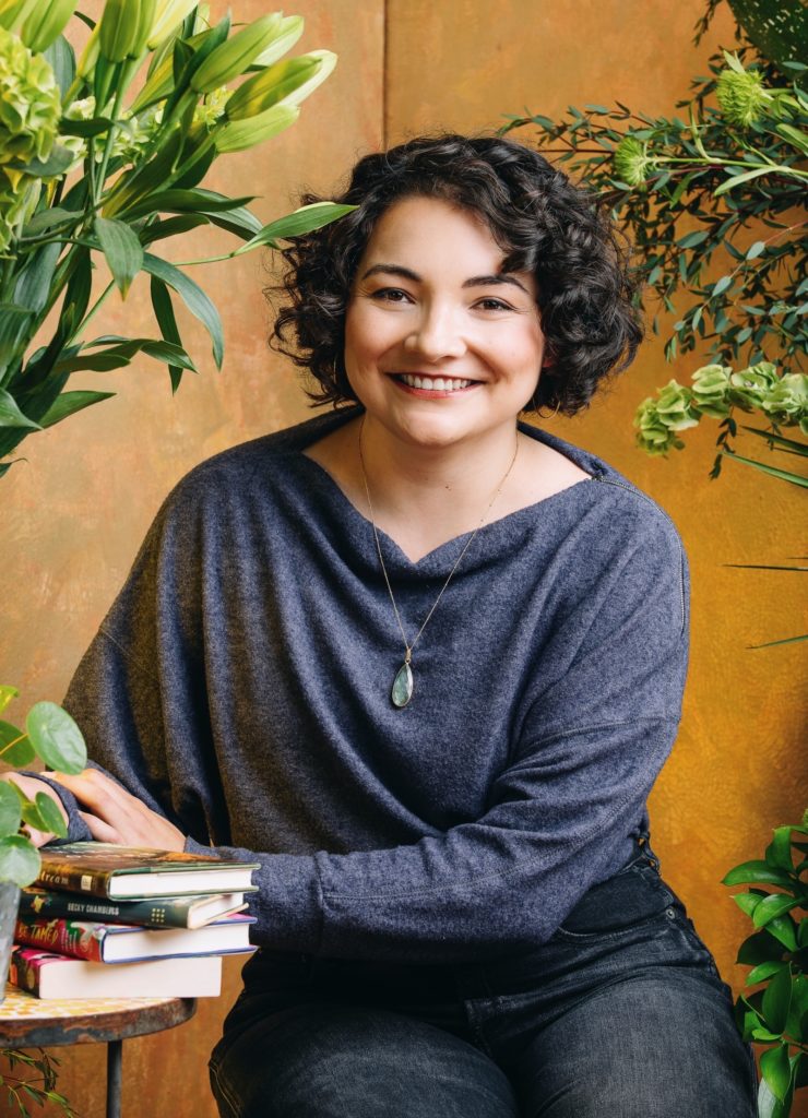 A photo of a light-skinned, female-presenting person with short, curly brown hair who is seated and smiling. Next to the figure, a small stack of books is visible on a side table, and green plants frame the foreground against an orange-gold backdrop.
